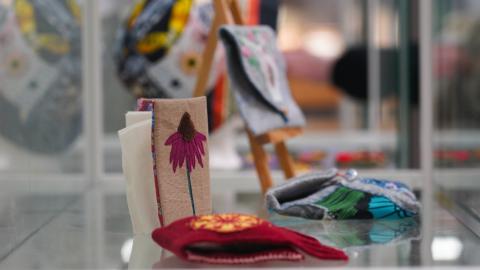 A display case featuring embroidery works. A floral-themed piece in the foreground.