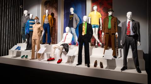 Miesten eri aikakauden vaatteisiin puetut mallinuket korokkeella näyttelytilassa.  
