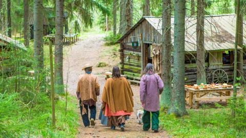 Kolme henkilöä kävelee metsätiellä selät kohti kameraa. Tien varralle on puisia rakennuksia.