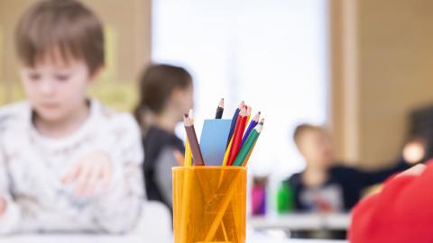 Kynäkuppi on pöydällä etualalla ja taustalla pieniä koululaisia.