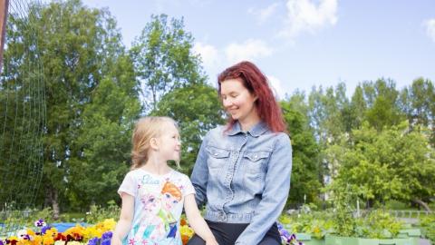 Mother and child look into each other's eyes and smile. Behind them you can see a forest of leaves and planting boxes with colourful pansies, as well as other plants.