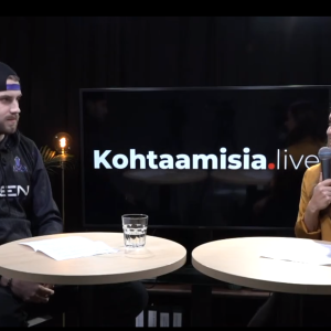 Nainen mikrofonin kanssa haastattelee miestä kahden pyöreän pöydän äärellä