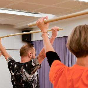 Kolme henkilöä keppijumppaa liikuntasalissa. Ylös nostetut kädet pitelevät puista keppiä.