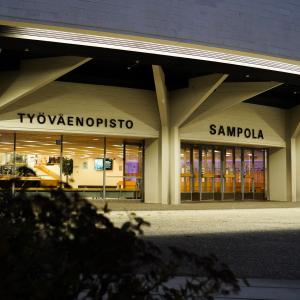 Sampola building in the dark. The wall is illuminated. There is a bush in the foreground.
