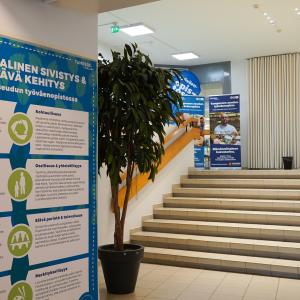 Seinällä juliste, jossa on paljon tekstiä. Taustalla huonekasvi ja portaikko.