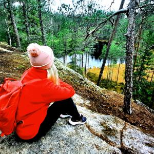 A girl in a red jacket with a rucksack on her back is sitting on a rock, looking out over a landscape with a forest and a small lake.