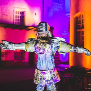 A metal statue of a human with their arms outstretched. The statue is wearing a helmet. In the background of the work you can see walls illuminated by colourful lights.