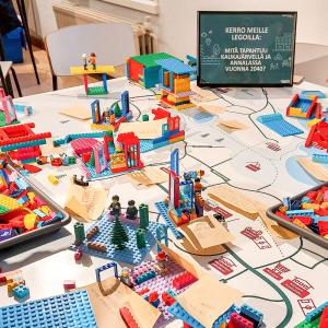 On a large table, a pile of colourful Lego blocks is being used by people to build the neighbourhood of their dreams on a map.