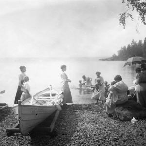 An old photograph showing people on the shore of a lake. Boats in the foreground and trees on the edge.