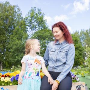 Mother and child look into each other's eyes and smile. Behind them you can see a forest of leaves and planting boxes with colourful pansies, as well as other plants.