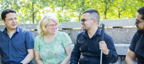 Four people are sitting on a bench in a summer landscape, looking at each other, with the trees in the background.
