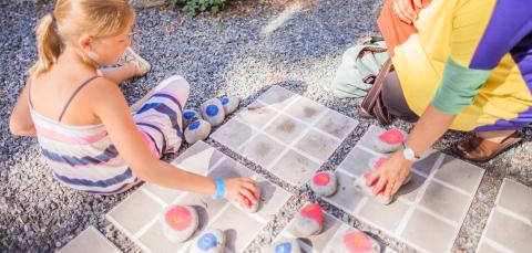 Lapsi leikkii ulkona kolmen suora -peliä erivärisillä kivillä aikuisen kanssa.