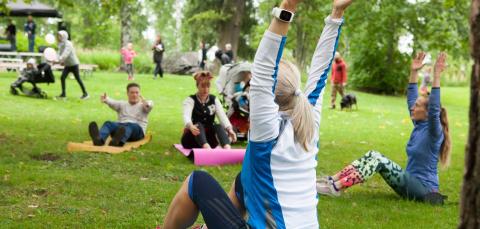 Ihmisiä puistossa jumppaamassa liikunnanohjaajan ohjeiden mukaan.