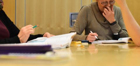 Henkilö lukee kirjaa ja nauraa. Edessä toisen opiskelijan käsi.