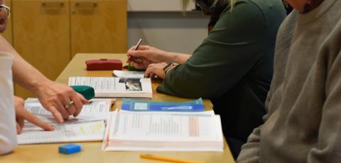 Henkilöt opiskelevat pöydän ääressä. Kirjat avoinna pöydällä.