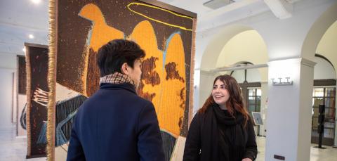 In the large gallery space, floor-to-ceiling murals hang from the ceiling. In front of them, two people are chatting, one with his back to the viewer, the other smiling at a friend.