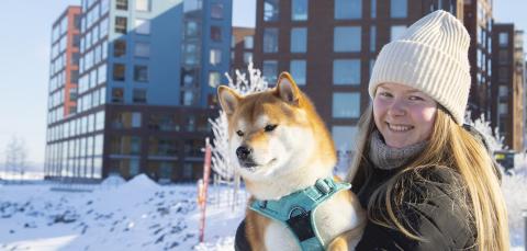 A person smiles and holds a brown and white dog on her lap in a winter landscape with apartment buildings in the background.