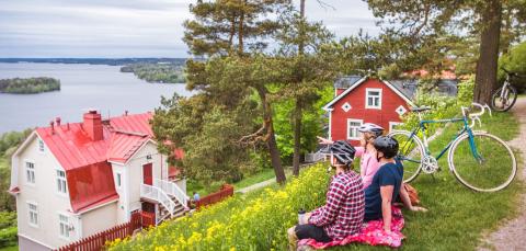 Three people with bicycle helmets on look out over Lake Pyhäjärvi on Pispala Esker, with wooden houses and bicycles in the landscape.