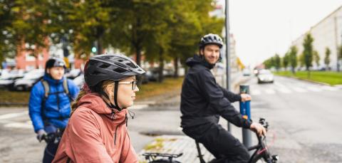 Kolme pyöräilijää kypärät päässä odottaa suojatien edessä valojen vaihtumista.