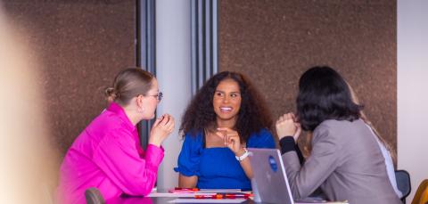 Three women are chatting at a table with an open laptop by their side.