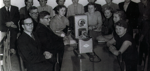 Joukko ihmisiä istuu epidiaskoopin ääressä.