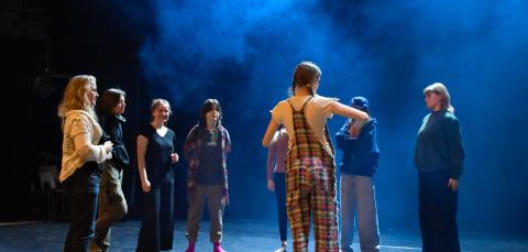 Seitsemän nuorta seisoo lavalla opettajan kanssa ringissä.