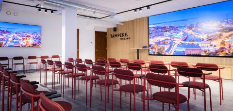 Kuvassa on Siluetin esitystila. Kuvan etualalla on neljä riviä punaisia penkkejä ja takana on vaalea, puinen seinä jolla on suuri näyttö. Näytössä on kuva Tampereen keskustasta.