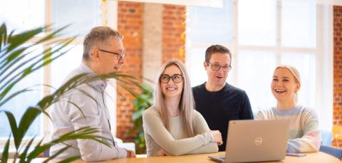 Four smiling people are standing at a desk in an office, chatting. There is a computer on the desk.