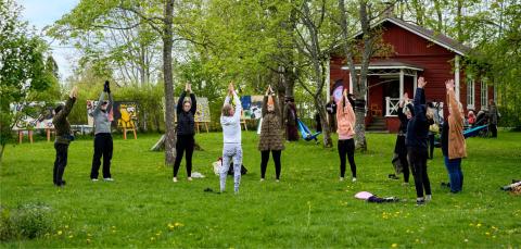 Iso joukko ihmisiä on ulkona jumppaamassa. He seisovat nurmikolla kädet ojennettuna korkealle ylös.