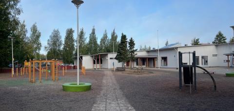 Metsäniityn päiväkodin ja koulun pihaa, taustalla näkyy rakennus. Piha on lähinnä avointa sorakenttää, jossa on puisia leikkivälineitä. on 