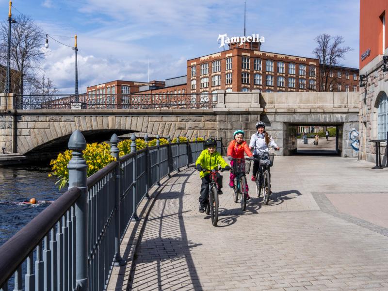 An adult and two children cycling in the spring landscape on the Valssipadonraitti. There are daffodil boxes on the bridge railing.