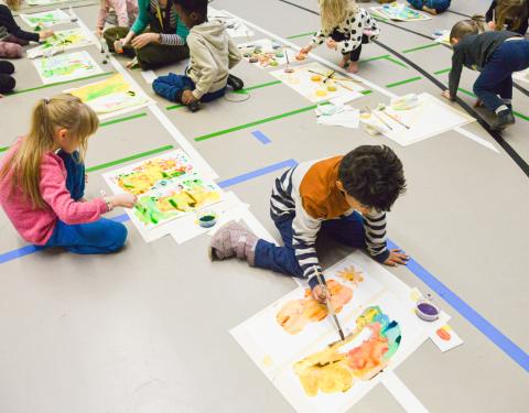 Lapsia maalaamassa vesiväreillä koulun jumppasalin lattialla.