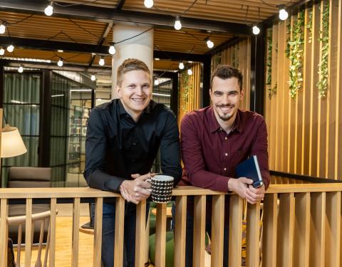 Kaksi miestä seisoo ja nojailee kaiteeseeen ja he katsovat hymyillen kohti., 