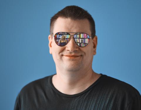 A middle-aged man looks toward the camera. His sunglasses reflect an image of the library.
