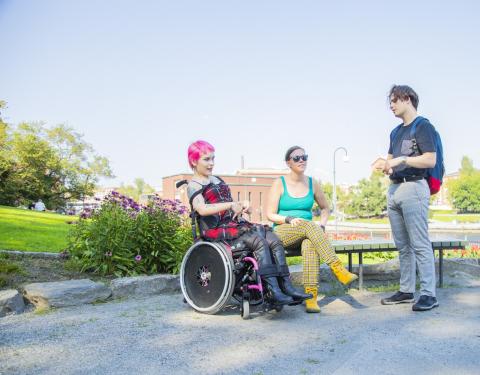 Pyörätuolia käyttävä pinkkitukkainen nainen istuu kesällä Koskipuistossa keskustelemassa kahden ystävänsä kanssa.