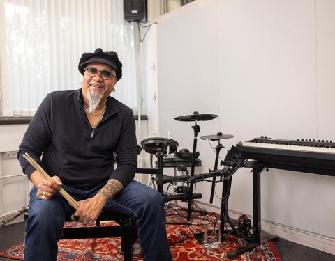 Mies istuu rumpusetin ja pianon vieressä rumpukapulat kädessään ja hymyilee kameralle