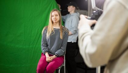 Opiskelija istuu tuolilla, taustalla vihreä kangas. Etualalla valokuvaaja ja takana opiskelija pitää kangasta.