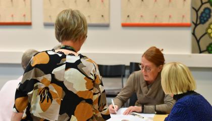 Henkilöt opiskelevat pöydän ääressä. Opettaja ohjaa vieressä.