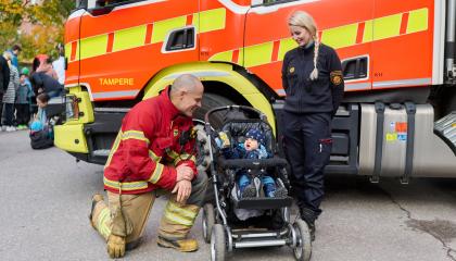 Kaksi aikuista seisoo ja yksi lapsi istuu rattaissa paloauton edessä.