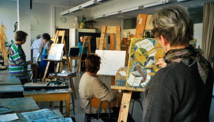 Henkilöitä maalaustelineiden edessä luokkahuoneessa. Pöydällä maalauksia.