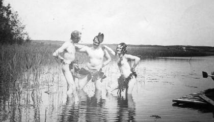 Three men stand naked in the water, frolicking with only their leaves for protection.