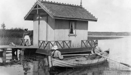 A boathouse, a boat and two people in the boat. One standing on the pier.