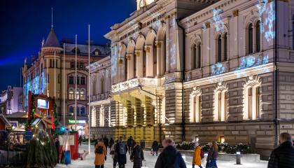 Raatihuone joulun aikaan valaistuna. Raatihuoneen edessä joulukuusien myyntipiste ja Ihmisiä kulkemassa ohitse. 