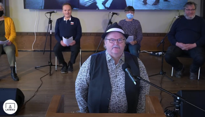 Hattupäinen mies seisoo ja laulaa. Taustalla neljä istuvaa taustalaulajaa.