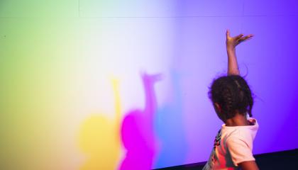 Lapsi käsi pystyssä, seinällä sateenkaaren värisiä varjoja.