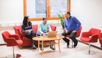 Yksi nainen ja kaksi miestä istuu toimistossa. Yksi heistä käyttää tietokonetta ja muut seuraavat työskentelyä