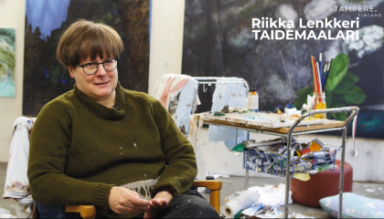 Nainen istuu ja taustalla on maalauksia ja maalaustarvikkeita