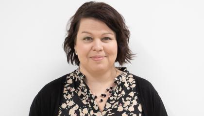 Hankkeen projektipäällikkö hymyilee kasvokuvassa. Tummat lyhyet hiukset, helmet kaulassa. 