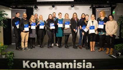 Iso ryhmä yhteisöagenteiksi valmistuneita henkilöitä seiso rivissä korokkeella. He pitävät käsissään valmennuksesta saatuja paperisia todistuksia. 