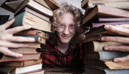 Kiharatukkainen nuori mies istuu ja nojaa samalla pöytään. Hän on kahmaissut syleilyynsä kaksi iso pinoa vanhoja kirjoja.  